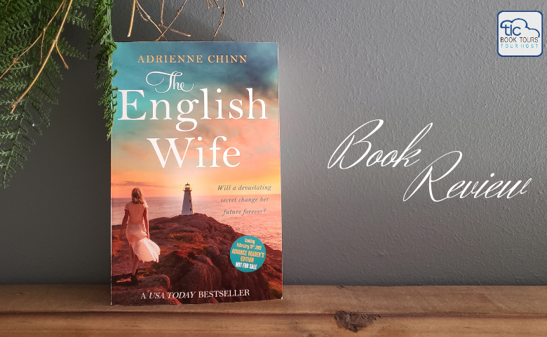 The English Wife book sitting on a wooden ledge in front of a slate gray/blue wall with greenery and branches just visible in the upper left corner.